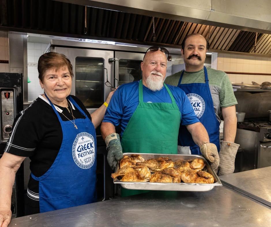 Three volunteers in the festival kitchen, two of them in Greek Festival aprons, holding out a tray of roast chickens.