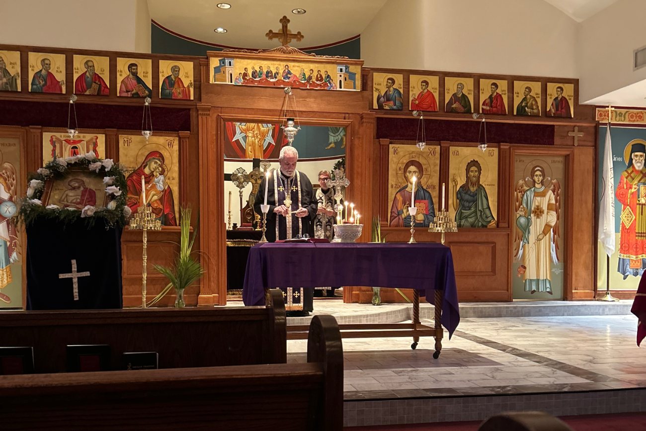 A priest reading by candlelight at the altar table in front of the iconostasis during a Holy Week service. At the left an icon stands draped in black and dressed with flowers.