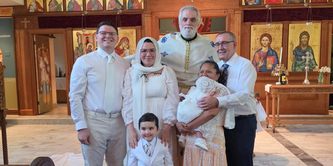 A family gathered with the priest in front of the iconostasis after a baptism, the newly baptised infant in a white gown.